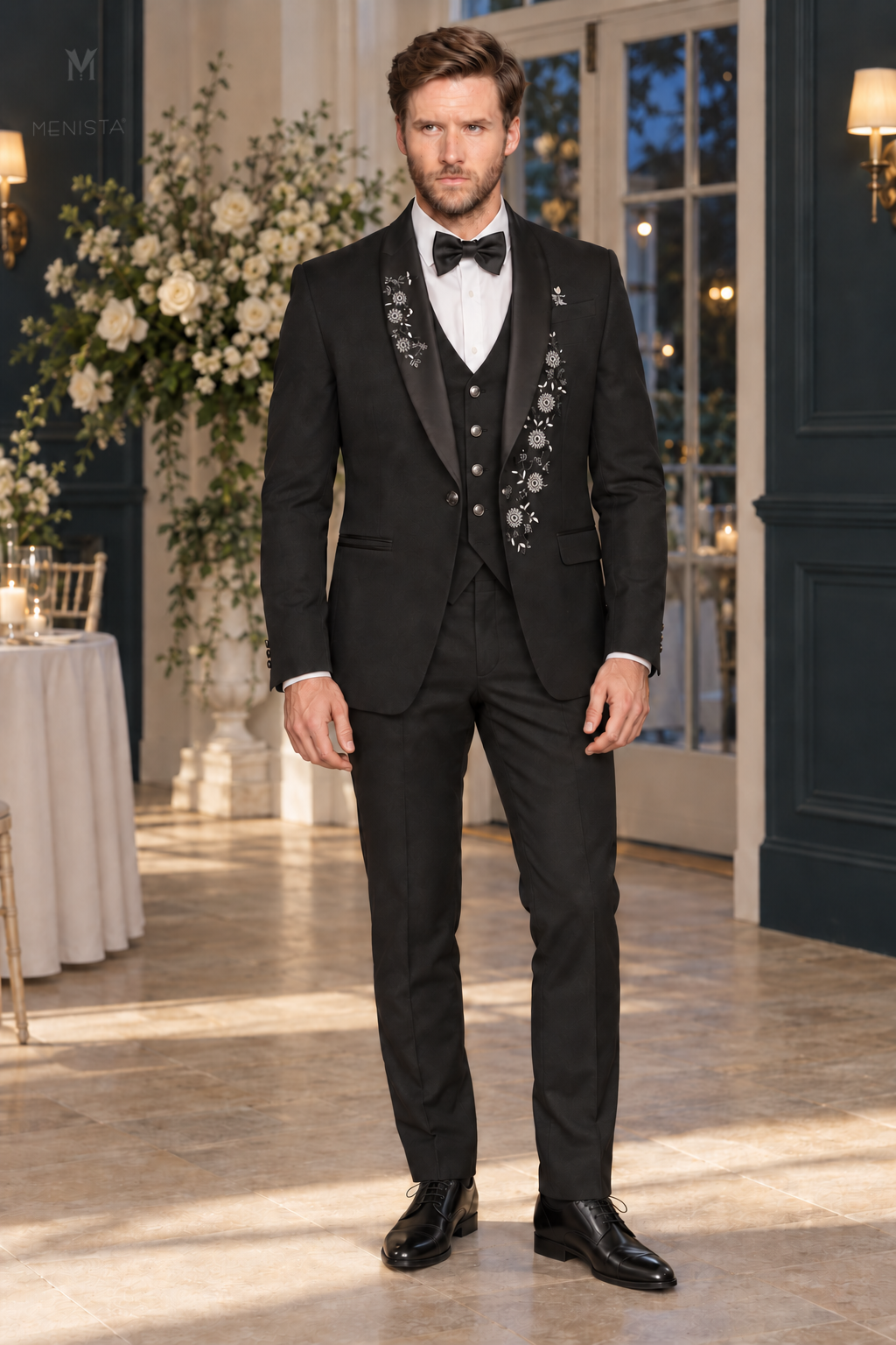 Jack wearing Menista Dorian black three-piece tuxedo with silver daisy floral embroidery and satin shawl lapels, full front view in a wedding orangery.