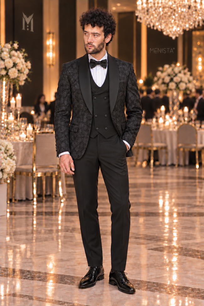Dorian black jacquard three-piece tuxedo on Michael, front full-body view, with plain black waistcoat and trousers in an indoor wedding venue.